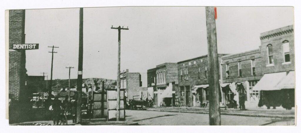A black-and-white photograph of North Greenwood Ave. in Tulsa, Oklahoma, prior to the 1921 Tulsa Race Riot. The photograph was taken looking north down the avenue from East Archer Street. The Williams Building is visible on the left side of the photograph. A sign that reads “Dentist” is visible on the second story of the Williams Building. Across the street the east side of N. Greenwood Ave. is visible. On the far right side of the photograph is the Woods Building. The Dreamland Theater is at the center of the photograph, the last building visible on the east side of the street. Several cars are parked on the east side of the avenue and people are seen walking or standing around the street. One figure on the corner outside the Williams building has a bicycle. There are no marks or inscriptions, front or back.