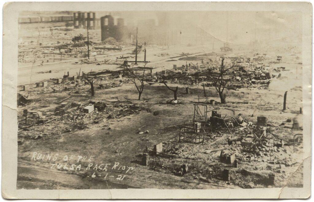 A sepia-toned photographic postcard depicting the destroyed Greenwood district of Tulsa, Oklahoma after the Tulsa Race Massacre. Taken from an elevated position, the image shows blocks of what had been homes and businesses reduced to rubble. In the right foreground several charred metal bedsteads are visible. A line of charred trees runs across the middle of the image. In the top left, across a wide avenue, stand a few empty brick building facades. At the bottom left corner, written into the negative and appearing as white text is [RUINS OF THE / TULSA RACE RIOT / 6-1-21]. 